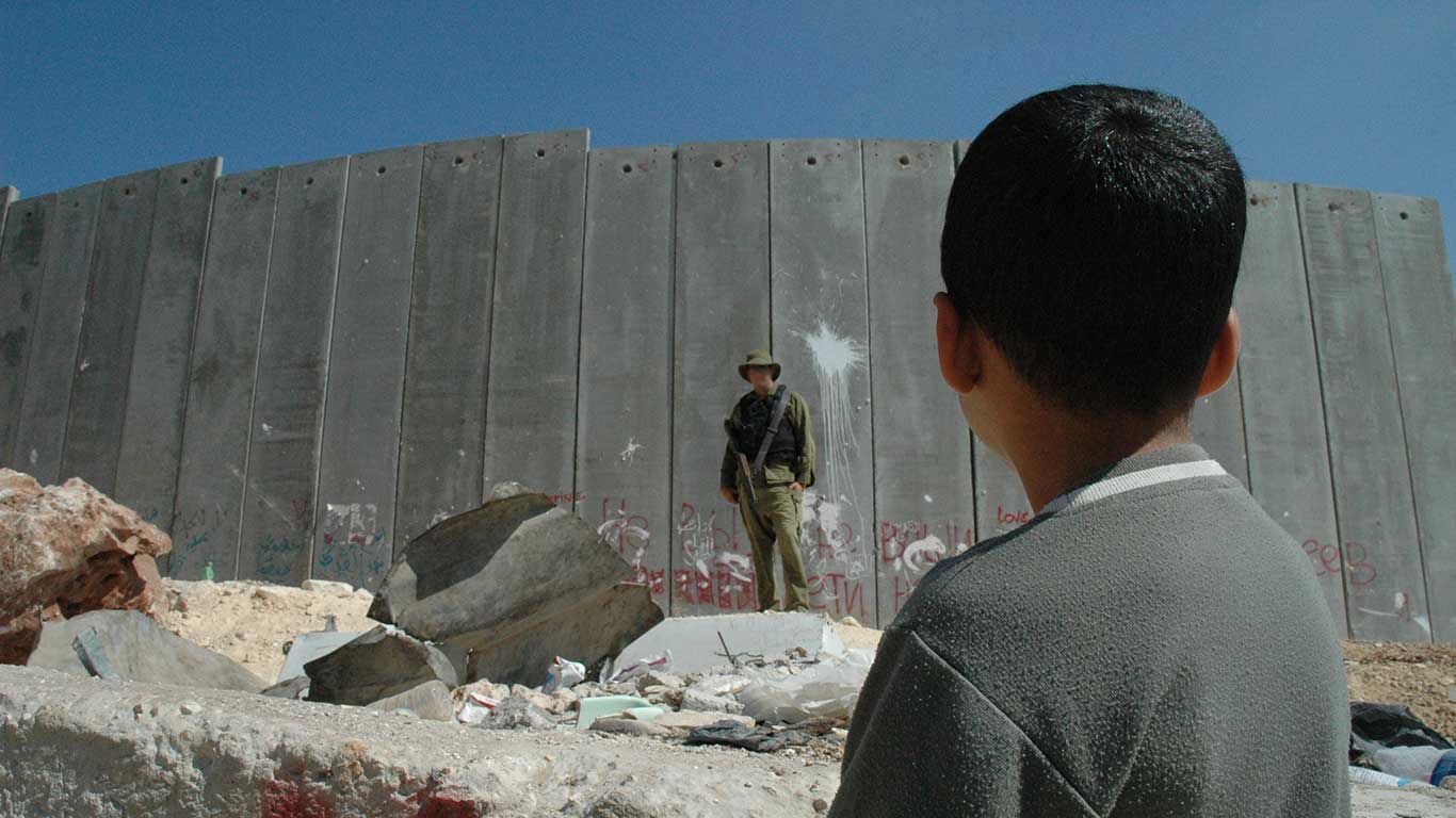 En palestinsk pojke och en israelisk soldat vid en tidigare konstruerad barriär på Västbanken. Bilden är från 2004. Foto: Justin McIntosh via Wikimedia Commons (CC BY 2.0).